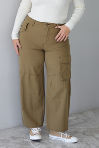 A closeup of a woman wearing olive khaki pants with a mid-rise waistline, pocket details, with a wide leg design, standing indoors against a white wall with white marble flooring. 