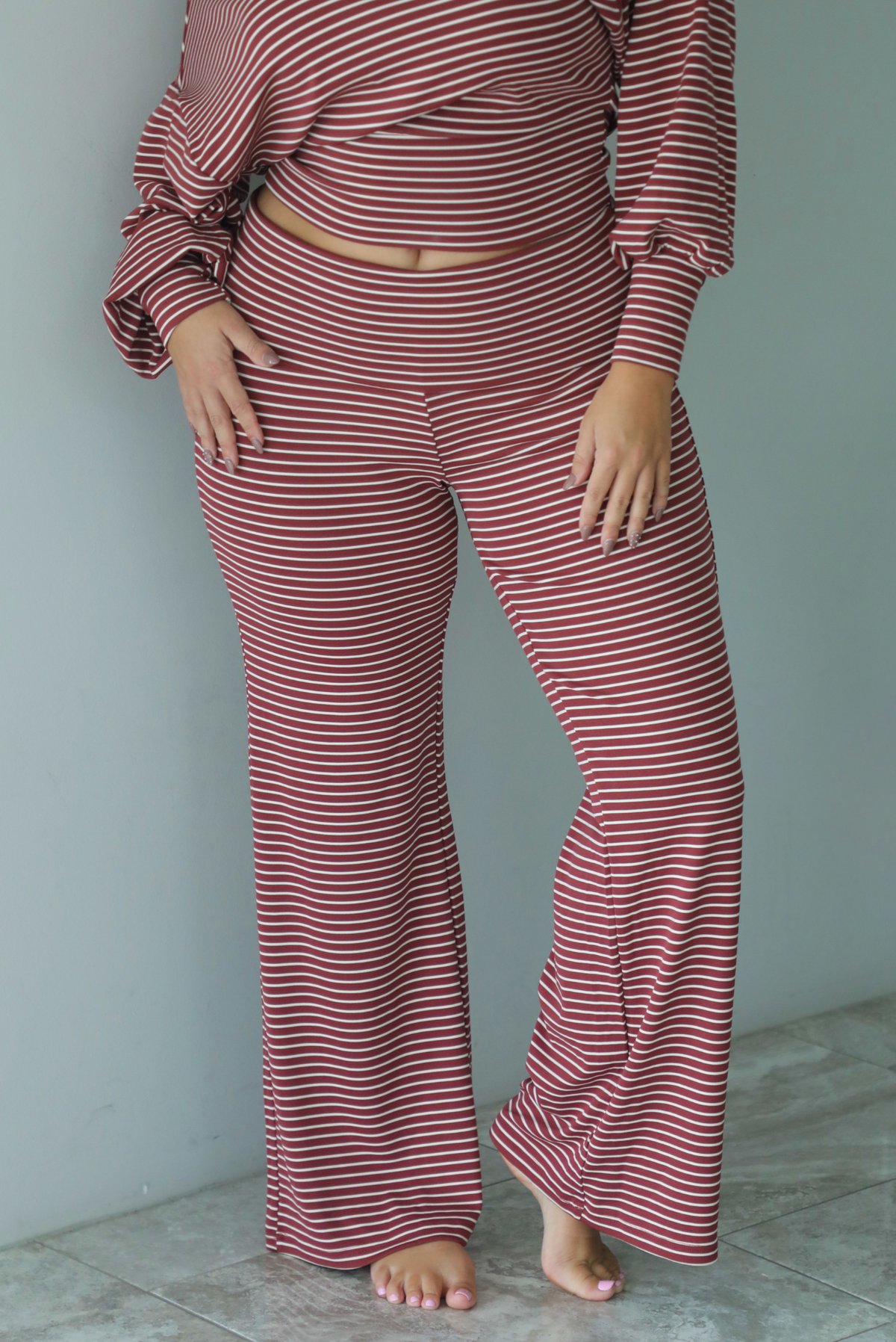 A woman wearing a maroon and white striped two piece matching set featuring a slouchy off the shoulder long sleeved top and mid-rise, folded over waistline wide leg pants, standing indoors against a white wall with white marble flooring. The photo shows a close up of the pants. 