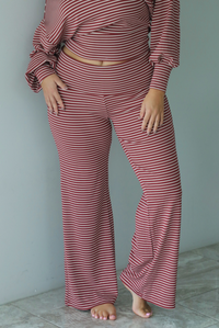 A woman wearing a maroon and white striped two piece matching set featuring a slouchy off the shoulder long sleeved top and mid-rise, folded over waistline wide leg pants, standing indoors against a white wall with white marble flooring. The photo shows a close up of the pants. 