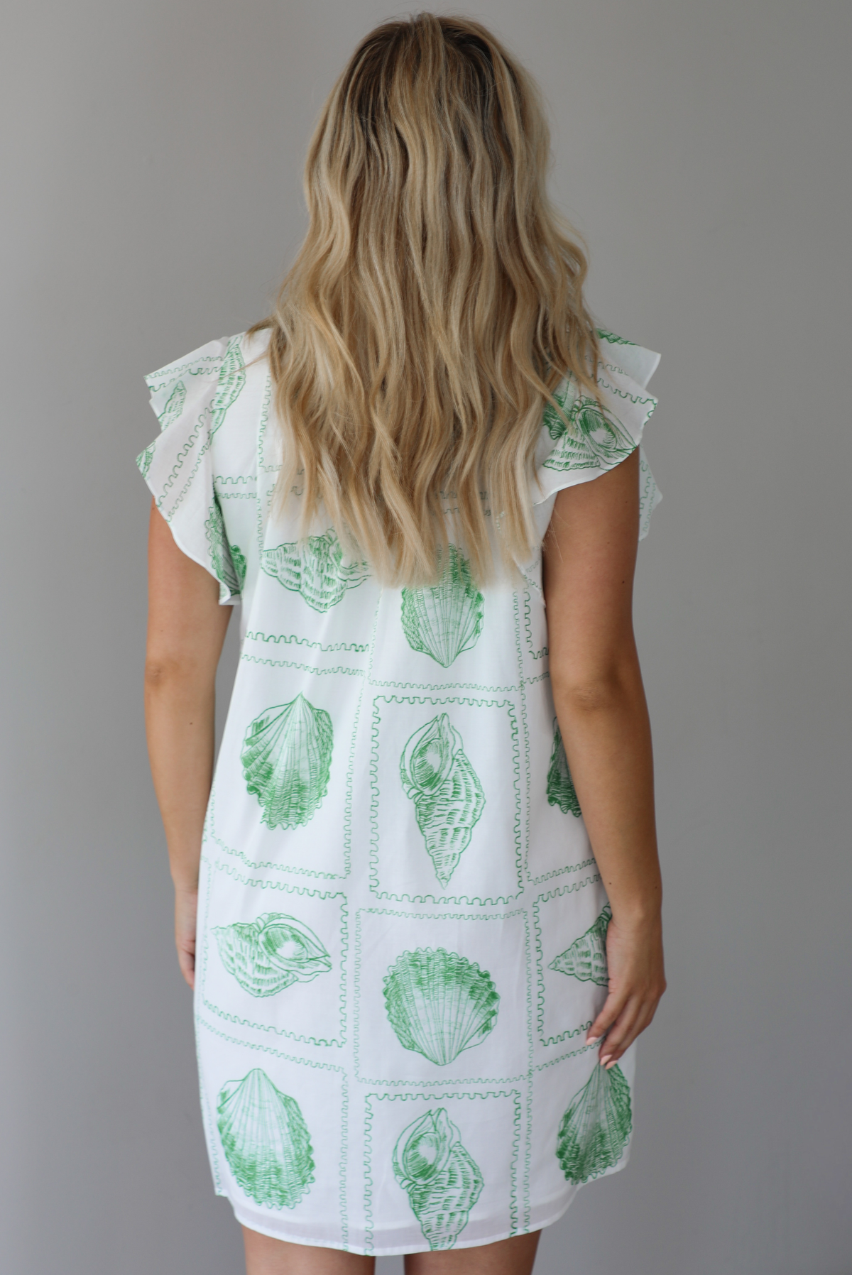 Woman wearing a white and green shell patterned dress with ruffled sleeves, functional pockets on either side, with a flowy skirt indoors against a white wall. The photo shows a closeup of the back of the dress. 