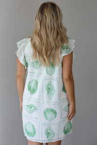 Woman wearing a white and green shell patterned dress with ruffled sleeves, functional pockets on either side, with a flowy skirt indoors against a white wall. The photo shows a closeup of the back of the dress. 