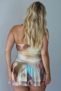 Woman wearing a multi-colored strapless mini dress that has a fitted bodice, a tie detail at the waistline, and a ruffled, flowy mini skirt indoors against a white wall. The photo shows the back of the dress. 