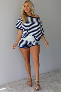 A woman wearing a navy and white horizontally striped off-the-shoulder top and matching striped shorts, standing indoors against a white wall with white marble flooring. 