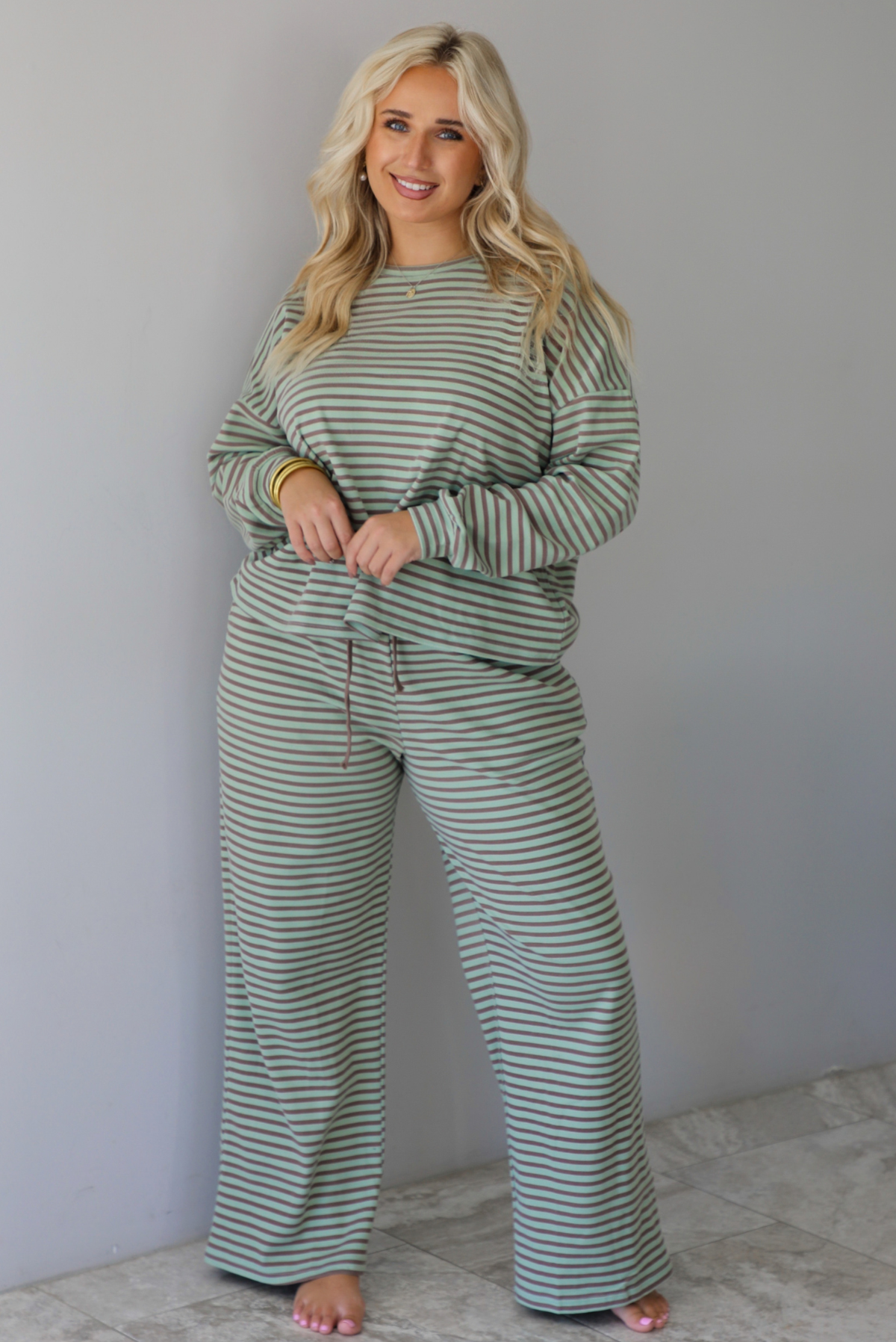 A woman wearing a pale green and brown striped two piece set featuring an oversized long sleeved top and high waisted, wide leg pants, standing indoors against a white wall with white marble flooring. 