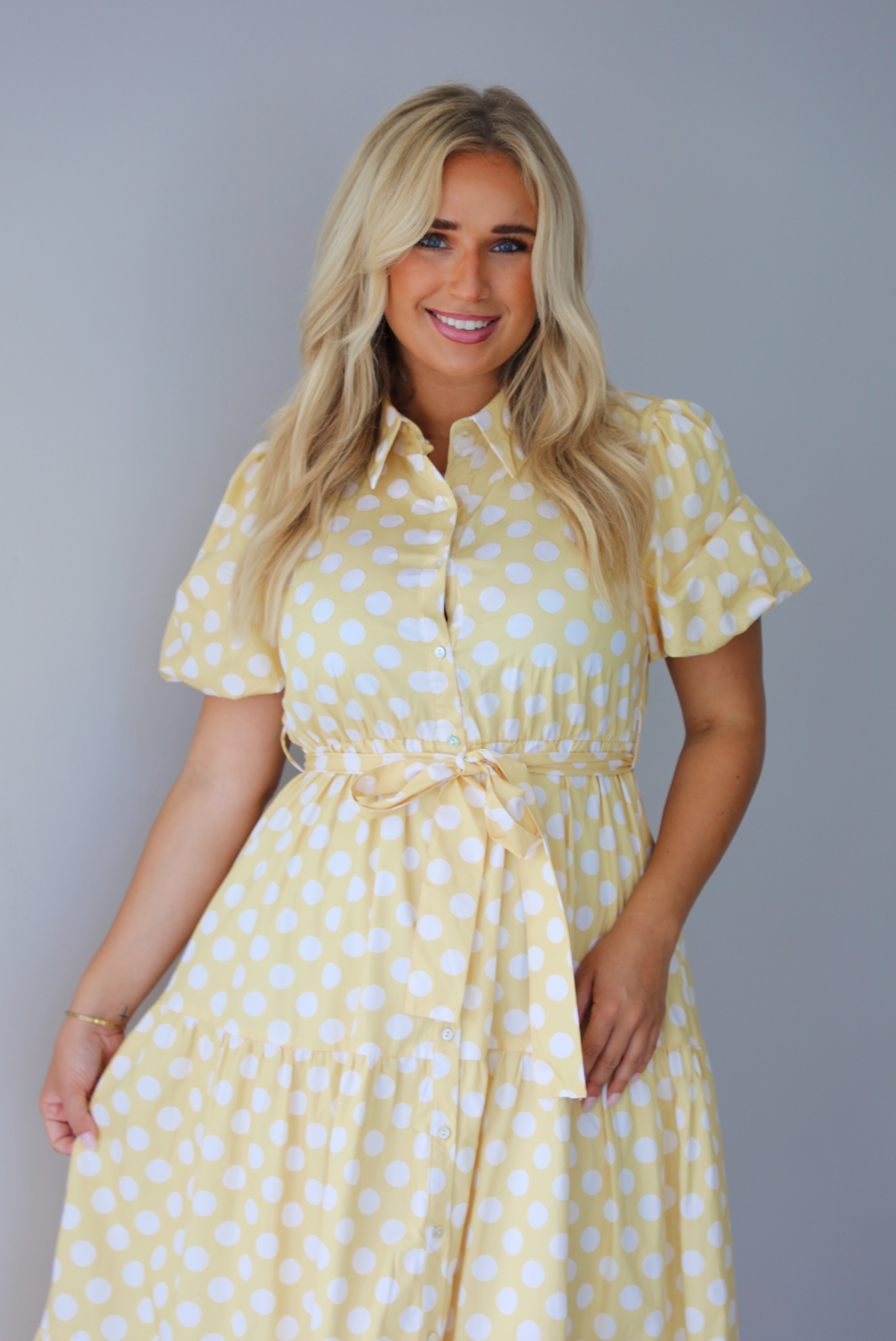 A woman wearing a yellow and white polka dot midi dress with puff sleeves and a tie waist and functional button closures along the front, standing indoors against a white wall.