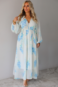 A woman modeling a white maxi dress with a blue floral print and a sheer, flowing overlay, standing indoors against a white wall with white marble flooring. 