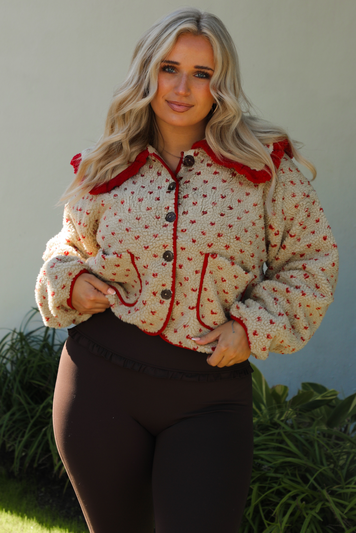 Woman model wearing a tan and red sherpa cardigan with a collared neckline and red trim with dark brown ahtleisure pants. The photo is taken outdoors with a white wall and greenery in the back. 