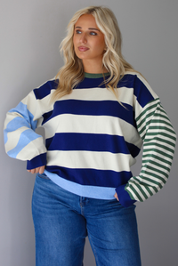 Woman wearing a navy, cream, blue, and green alternating striped design throughout, paired with dark denim jeans, standing indoors against a white wall.