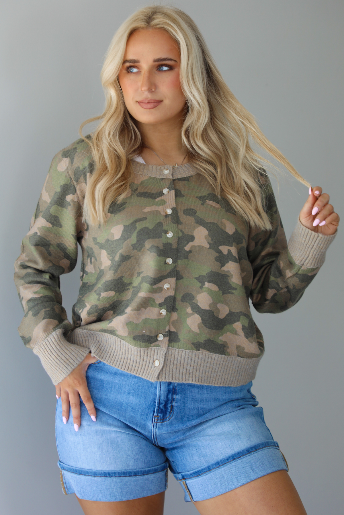 Woman wearing a camo cardigan with functional button closures in the front tucked into high waisted denim shorts. The photo is taken in front of a white wall.