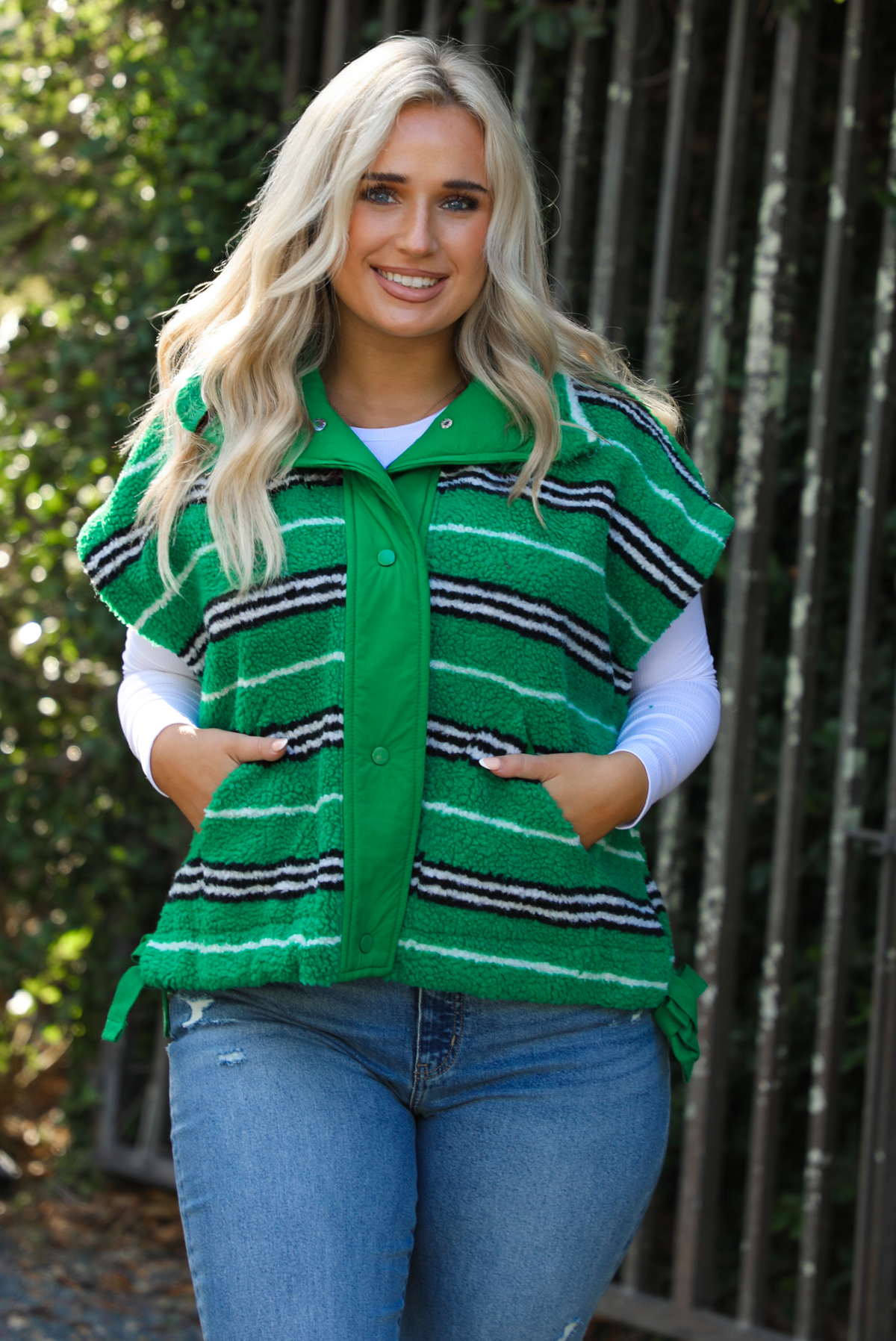 Woman model wearing a green, black, and white striped vest that has a sherpa texture to it, functional button closures in the front, and tie closures on either side, It is paired with a white long sleeved top, a medium wash pair of denim jeans, and tan ugg boots. The photo is taken outdoors with a metal gate and greenery in the back. The vest is shown close up, displaying the buttons closed and the pockets in use on either side.