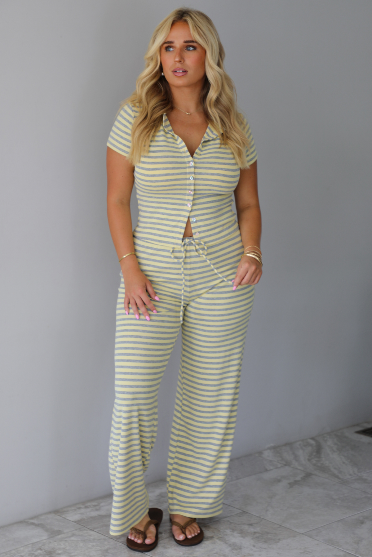 A woman wearing a two piece set consisting of a collared, button-down, short sleeve top and high waisted, drawstring, wide leg pants, standing indoors against a white wall with white marble flooring. 