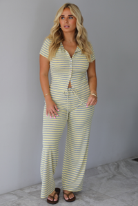 A woman wearing a two piece set consisting of a collared, button-down, short sleeve top and high waisted, drawstring, wide leg pants, standing indoors against a white wall with white marble flooring. 