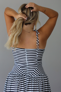 A woman viewed from behind wearing a sleeveless black and white horizontally striped maxi dress, focusing on the zipper closure and adjustable steps in the back, standing indoors against a white wall. 
