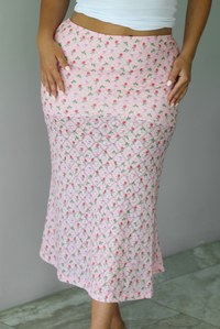 Woman wearing a pink midi length skirt with a multi-colored floral and bow repeating pattern indoors against a white wall with white marble flooring. The photo shows the front of the skirt. 