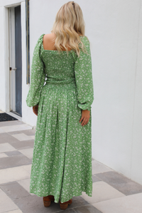 Woman model wearing a green maxi length dress that has a white floral print pattern, long sleeves, smocked bodice, and a flowy skirt. The model is holding the skirt up on one side. The photo is taken outdoors against a white wall.  The photo shows the back of the dress. 