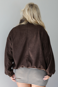 A woman shown from behind wearing a dark brown suede jacket over a dark brown, light blue, and cream striped mini dress, standing indoors against a white wall.