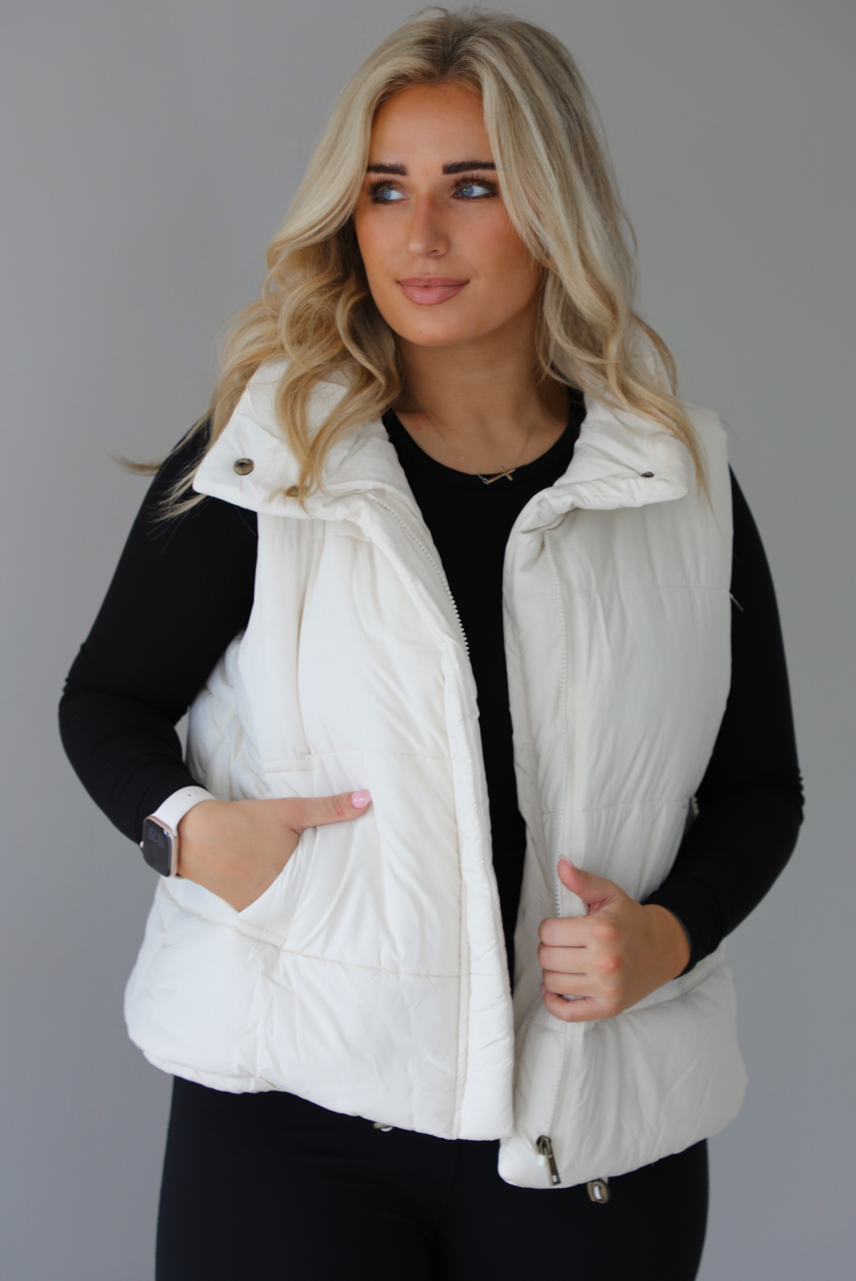 Woman wearing a light sand colored puffer vest with a fitted black long sleeved top and fitted leggings. The vest has functional button closures at the top, a zipper closure in the front, and functional pockets on either side. The photo shows the vest unzipped. The photo is taken indoors against a white wall with white marble flooring. 