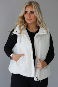 Woman wearing a light sand colored puffer vest with a fitted black long sleeved top and fitted leggings. The vest has functional button closures at the top, a zipper closure in the front, and functional pockets on either side. The photo shows the vest unzipped. The photo is taken indoors against a white wall with white marble flooring. 