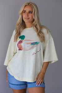 Woman wearing an off-white t-shirt with a graphic design of a mallard duck on the front with blue denim shorts. The photo is taken indoors against a white wall.