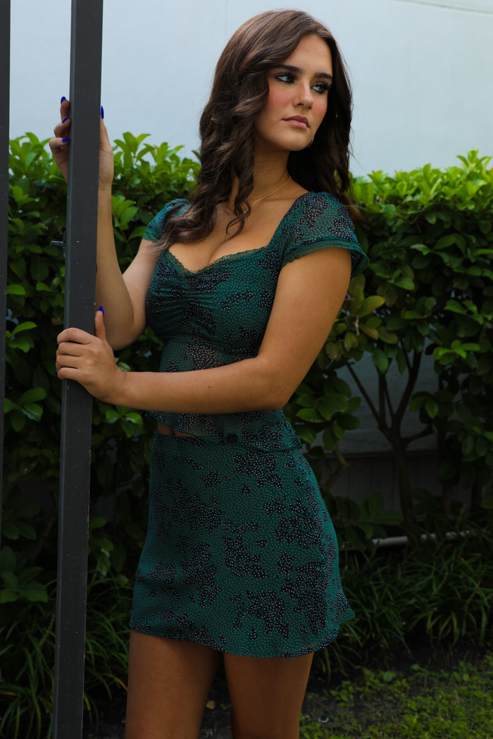 Woman model wearing a green with black and white pattern throughout set featuring a cropped sheer mesh top and mini skirt. The set is paired with tall black boots that stop just under the knee and is taken outdoors with greenery in the back. The photo shows a close up of the cinched detail at the bust of the top and the lace detailing at the short sleeve hem.