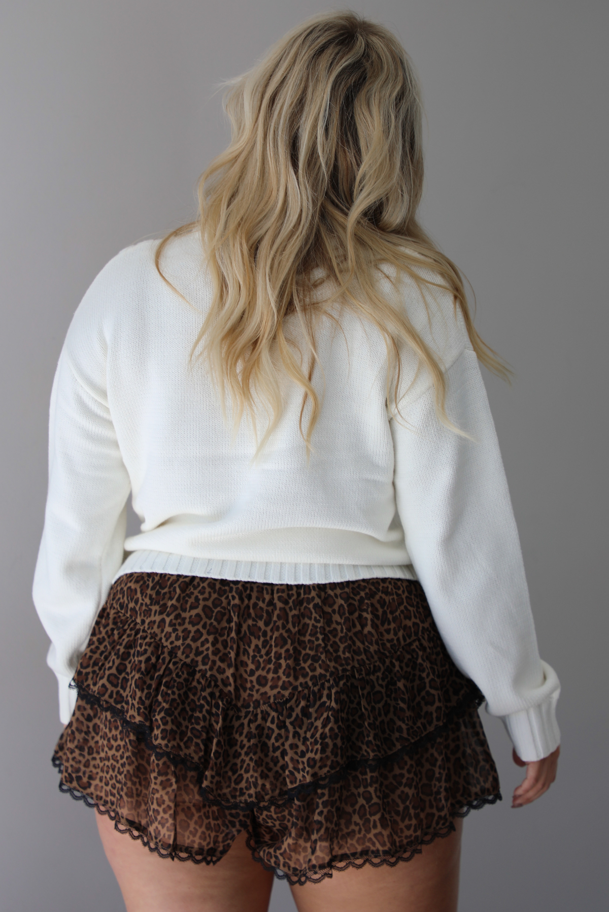 A woman shown from behind wearing a white sweater with bow details along the front, untucked to show the full length, paired with black and brown leopard print frilly shorts with black lace trim detailing, standing indoors against a white wall.