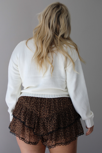 A woman shown from behind wearing a white sweater with bow details along the front, untucked to show the full length, paired with black and brown leopard print frilly shorts with black lace trim detailing, standing indoors against a white wall.