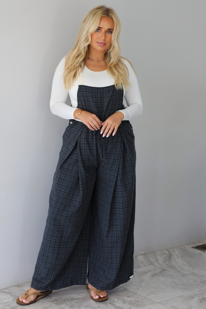 A woman modeling a white long-sleeve top and dark gray plaid wide-leg overalls, standing indoors against a white wall with white marble flooring. 