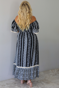 A woman shown from behind wearing a black and white maxi dress with a white floral print that alternates between a cluster appearance and a striped appearance, with off the shoulder sleeves and a flowy skirt, standing indoors against a white wall with white marble flooring. 