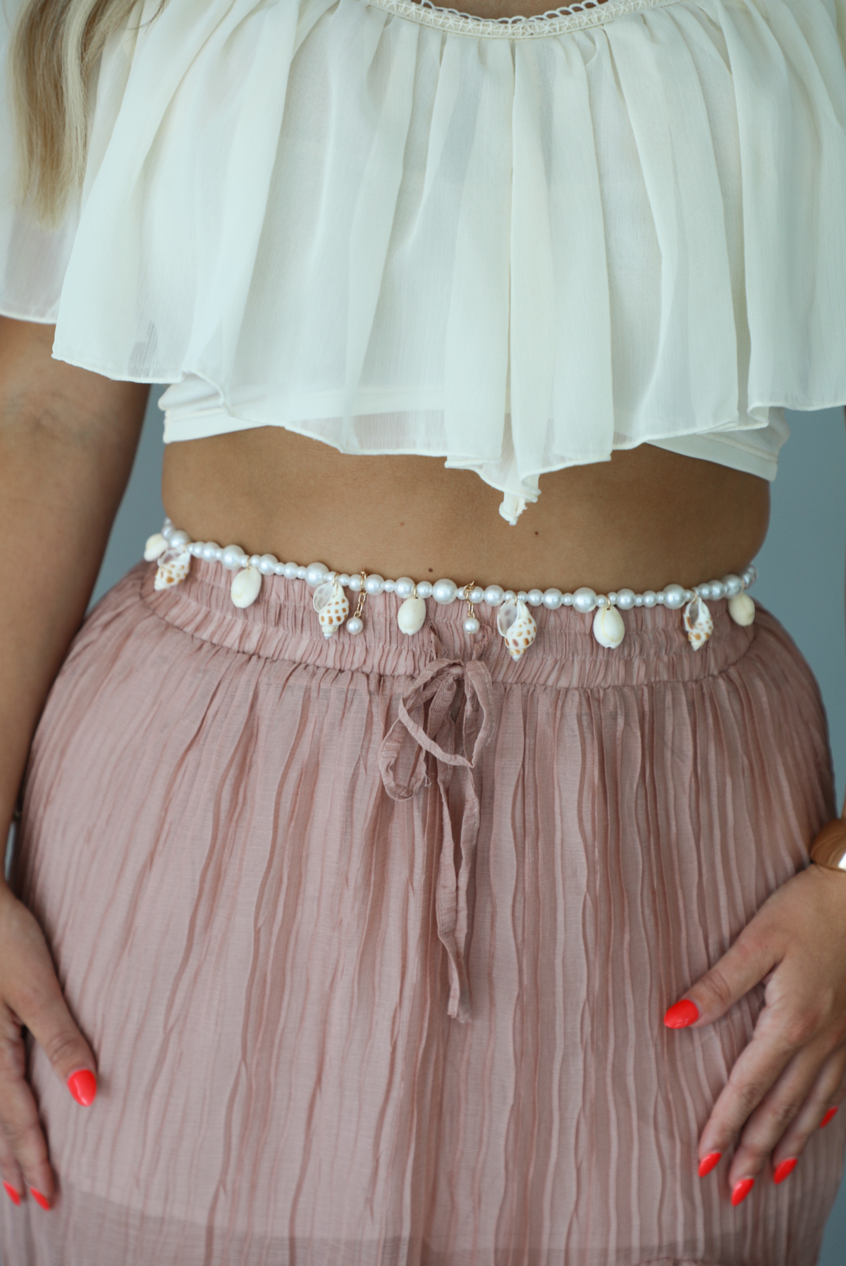 Closeup of a woman wearing a cream cropped top with a blush maxi skirt, focusing in on the pearl and shell belly chain belt. 