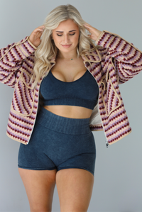 Woman wearing a pink, burgundy, lilac, and sand colored jacket with a charcoal colored two-piece set featuring a cropped sports bra and high waisted short biker shorts. The photo is taken indoors against a white wall.