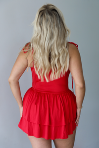 Woman wearing a red mini dress that has thin, spaghetti straps, a fitted bodice, and a flowy, ruffled skirt indoors against a white wall. The photo shows the back of the dress. 