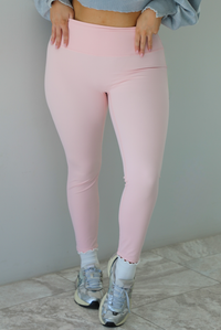 Woman wearing baby pink, fitted, high waisted leggings. The leggings are paired with white ankle socks and silver sneakers. The photo is taken indoors against a white wall with white marble flooring. 