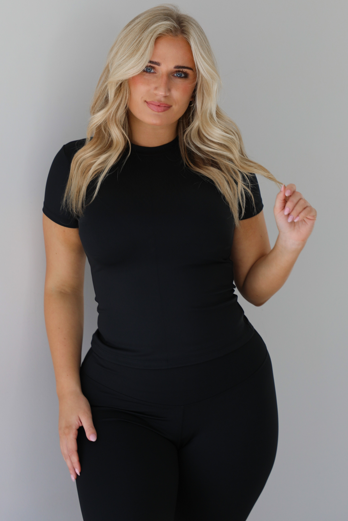 Woman wearing a fitted two piece black set featuring a short sleeved top and fitted leggings. The photo shows a closeup of the top of the set and the top of the pants. The photo is taken indoors against a white wall.