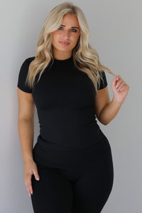 Woman wearing a fitted two piece black set featuring a short sleeved top and fitted leggings. The photo shows a closeup of the top of the set and the top of the pants. The photo is taken indoors against a white wall.
