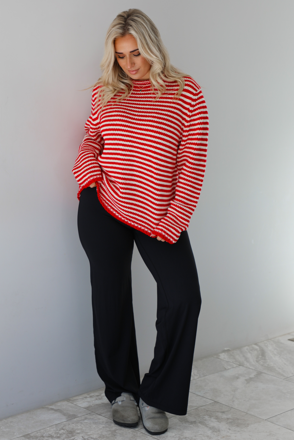 Woman wearing a white and red striped sweater that has a high neckline, an oversized fit, and is paired with black leggings. The photo is taken indoors against a white wall with white marble flooring. 