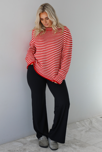 Woman wearing a white and red striped sweater that has a high neckline, an oversized fit, and is paired with black leggings. The photo is taken indoors against a white wall with white marble flooring. 