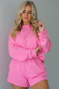 Woman wearing a hot pink two piece set featuring an oversized sweatshirt and high waisted shorts. The photo is taken indoors against a white wall and shows a closeup of the set.