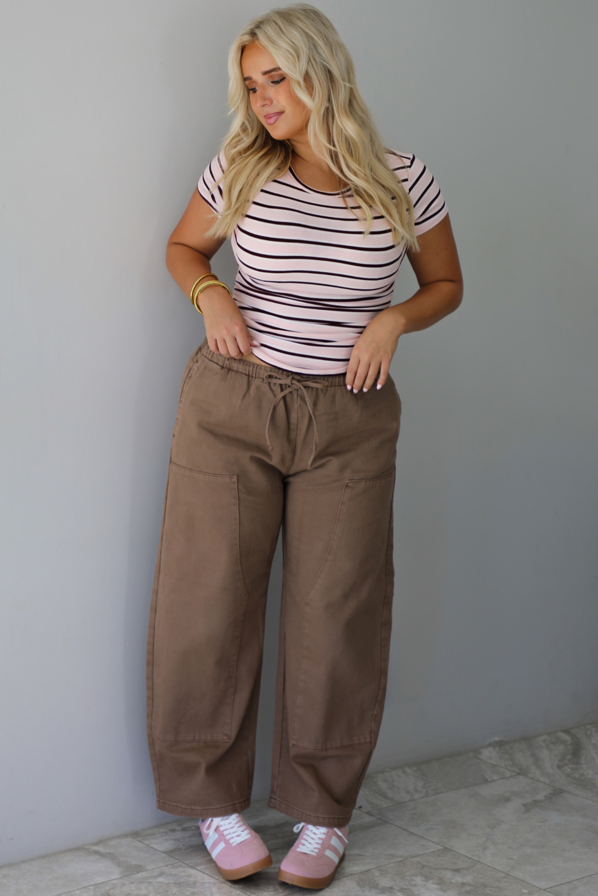 A woman wearing a pink baby tee that has a burgundy and white striped design throughout, paired with mocha barrel denim, standing indoors against a white wall with white marble flooring. 