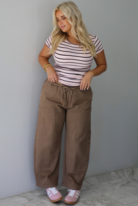 A woman wearing a pink baby tee that has a burgundy and white striped design throughout, paired with mocha barrel denim, standing indoors against a white wall with white marble flooring. 