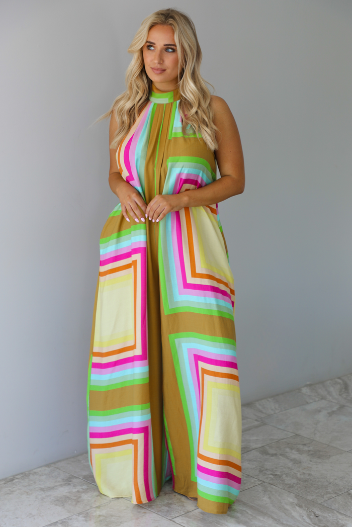 Woman wearing a colorful jumpsuit that has a high neckline, no sleeves, and a flowy cut with flared hem pants. The photo is taken indoors against a white wall with white marble flooring. 