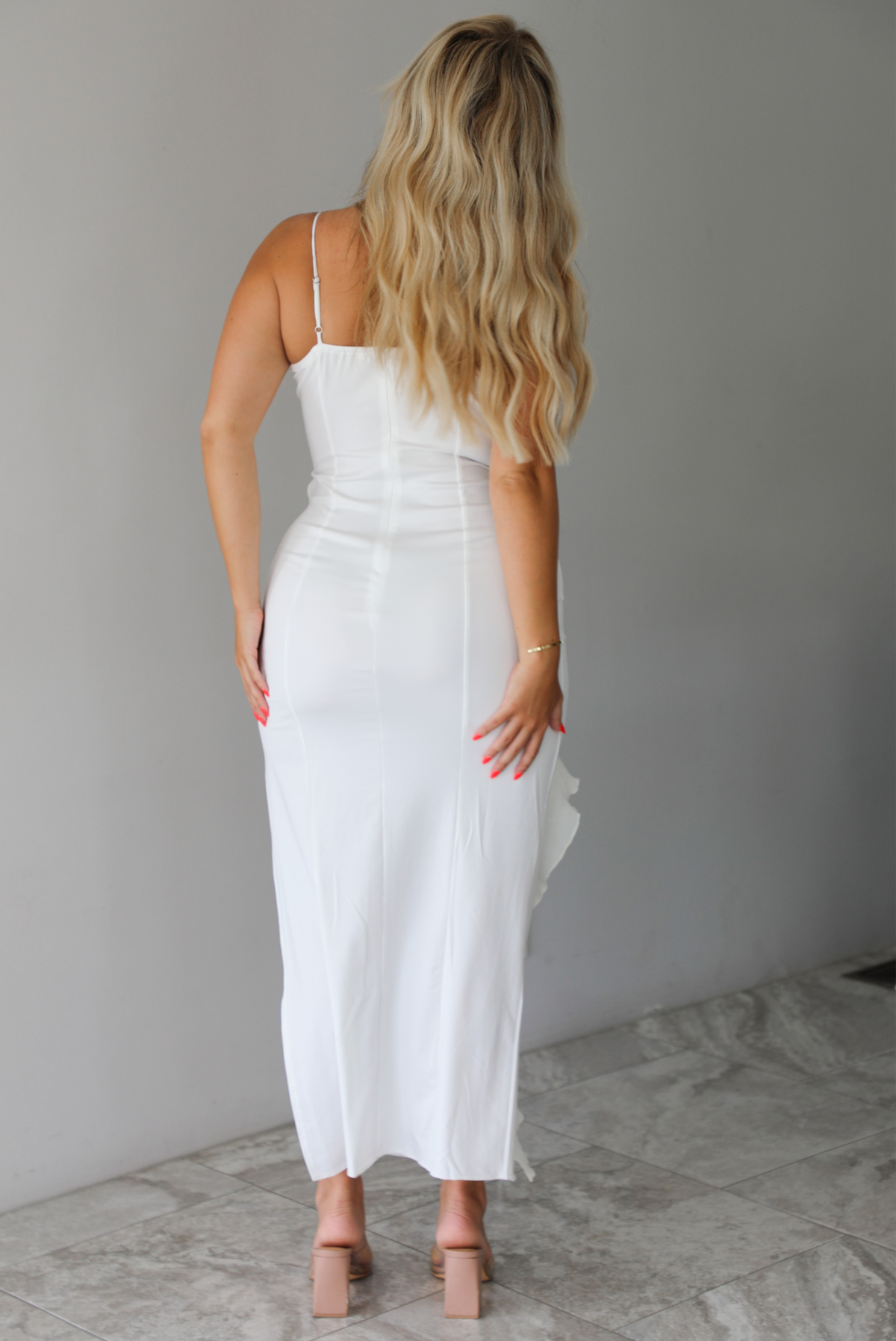 Woman wearing a white maxi length dress with ruffled details along the front indoors against a white wall with white marble flooring.  The photo shows the back of the dress, showing its adjustable straps and zipper closure. 