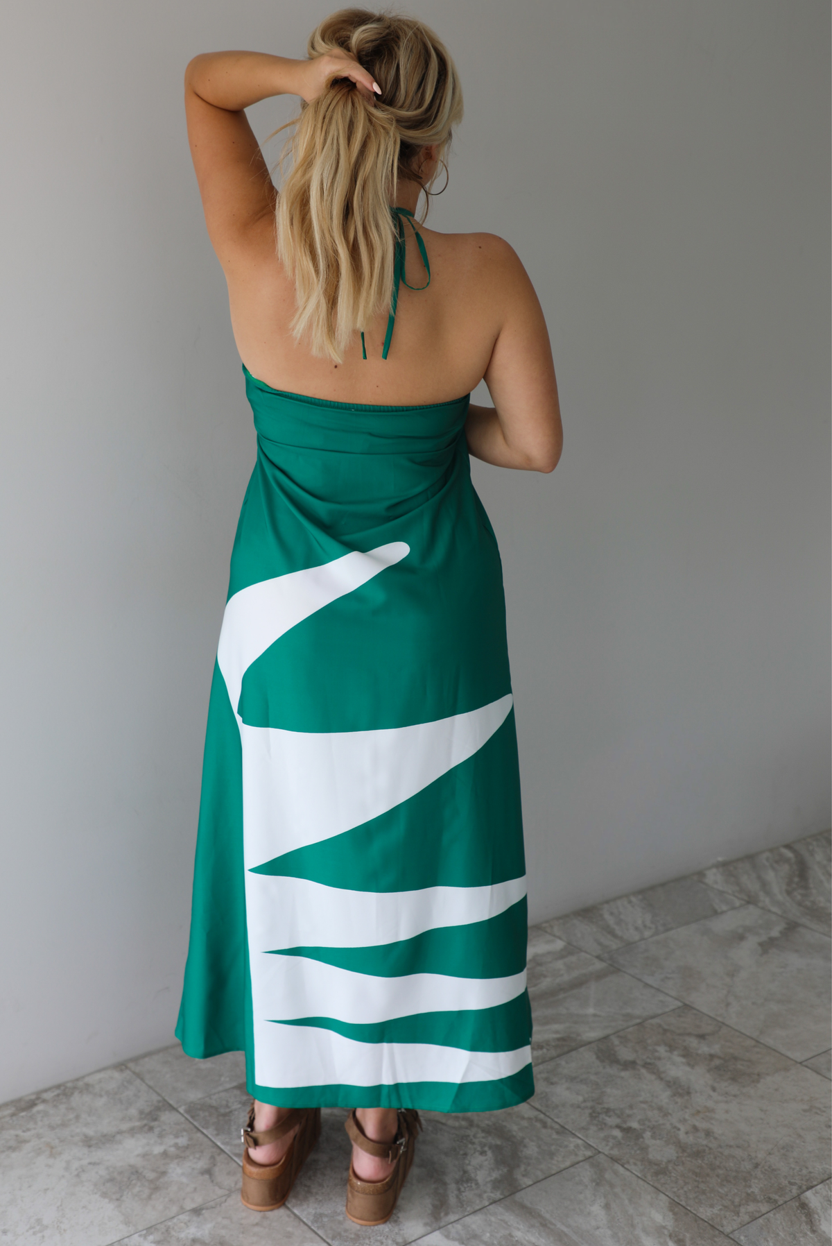 Woman wearing a green and white maxi dress with a halter neckline indoors against a white wall with white marble flooring. The photo shows the back of the dress. 
