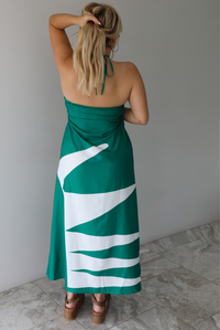 Woman wearing a green and white maxi dress with a halter neckline indoors against a white wall with white marble flooring. The photo shows the back of the dress. 