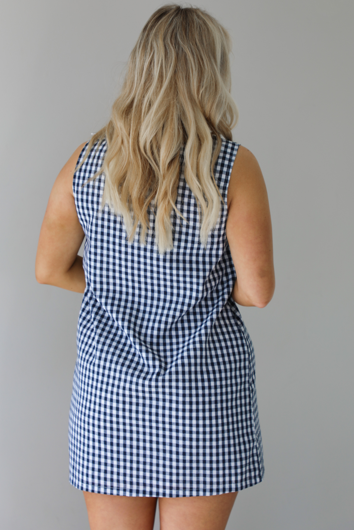 Woman wearing a navy and white gingham print patterned dress that has non-functional button closures on the left side. The photo is taken indoors against a white wall. The photo shows the back of the dress. 