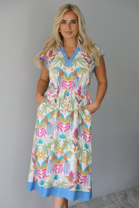 A woman wearing a short sleeved midi dress with a colorful tropical leaf print and light blue trim indoors against a white wall with white marble flooring. 