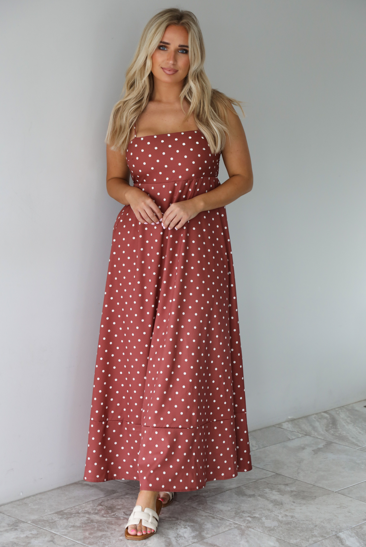 Woman wearing a spaghetti strap, maxi length dress that has a rust color throughout with a white polka dot pattern. The photo is taken indoors against a white wall with white marble flooring.