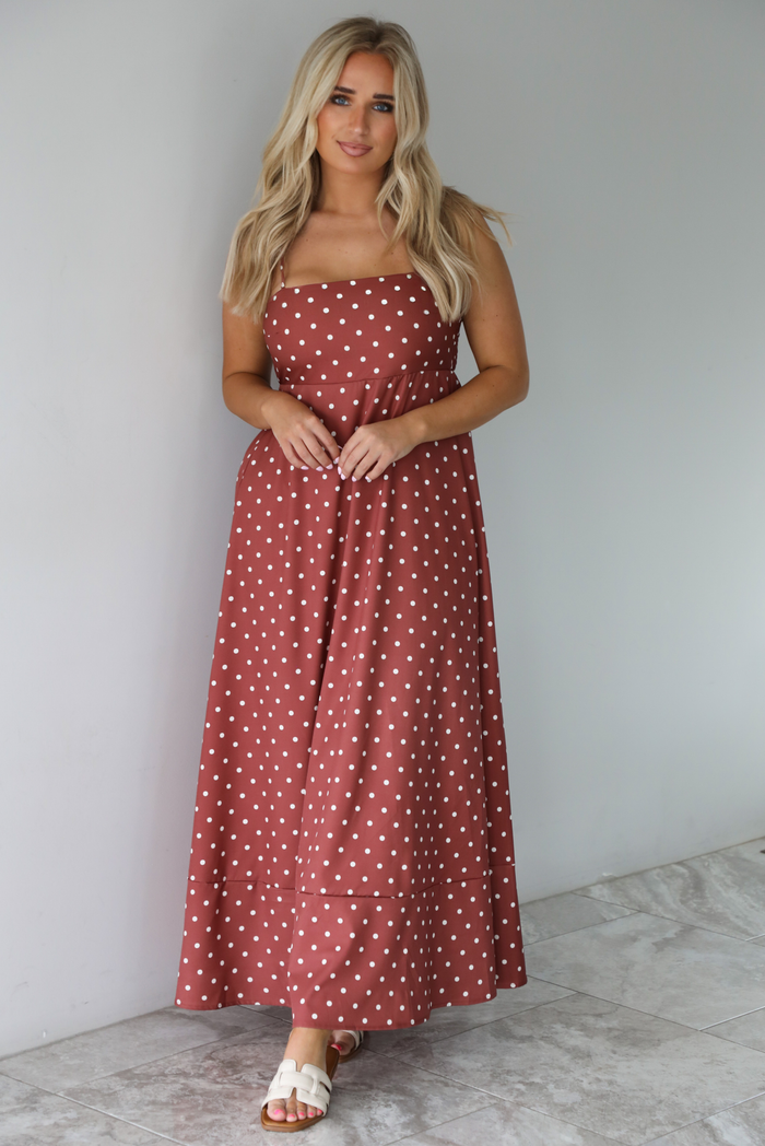 Woman wearing a spaghetti strap, maxi length dress that has a rust color throughout with a white polka dot pattern. The photo is taken indoors against a white wall with white marble flooring.