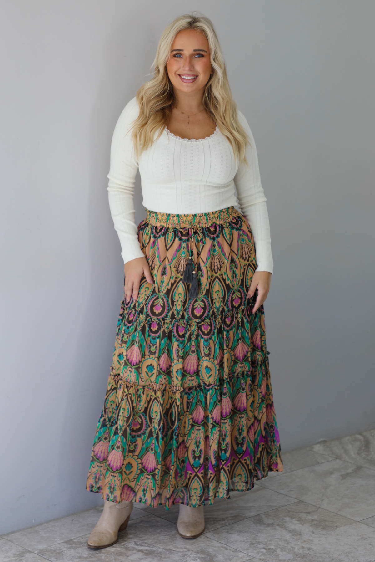 A woman wearing a long sleeved cream top and multi-colored maxi length skirt paired with knee high sand colored ankle boots, standing indoors against a white wall with white marble flooring. 