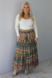 A woman wearing a long sleeved cream top and multi-colored maxi length skirt paired with knee high sand colored ankle boots, standing indoors against a white wall with white marble flooring. 
