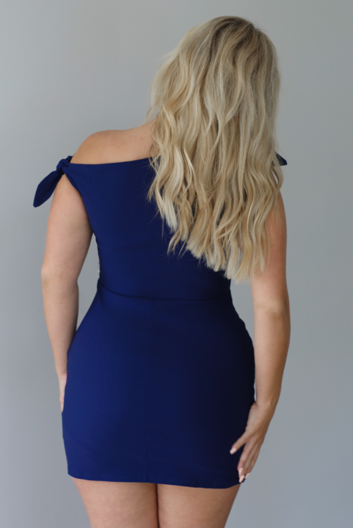 Woman wearing a navy mini dress with an off the shoulder design indoors against a white wall. The photo shows the back of the dress. 